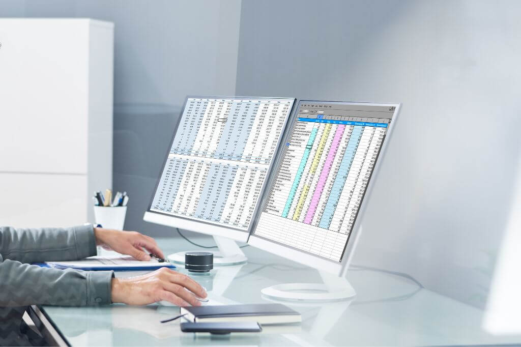Two screens on a desk containing data spreadsheets