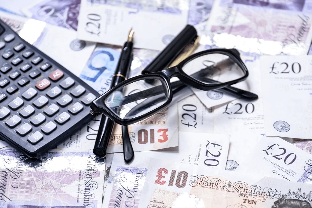 British pound notes scattered on a table with a calculator, pen and glasses