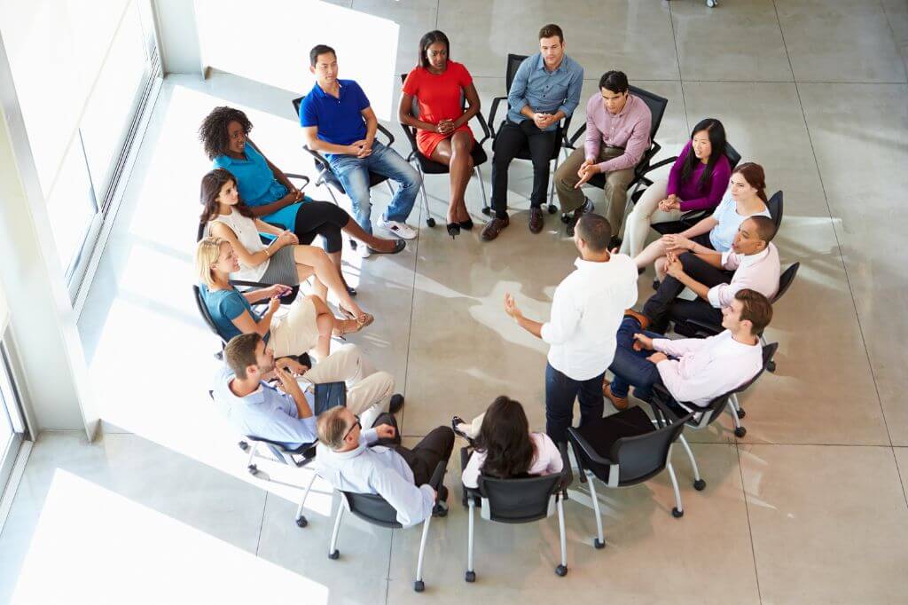 Office workers sat on chairs in a circle while one person is stood from his chair talking to the group.